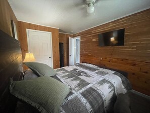 Camera da letto con parete in legno, un letto con coperta a motivi decorativi, una lampada e un televisore fissato al muro.