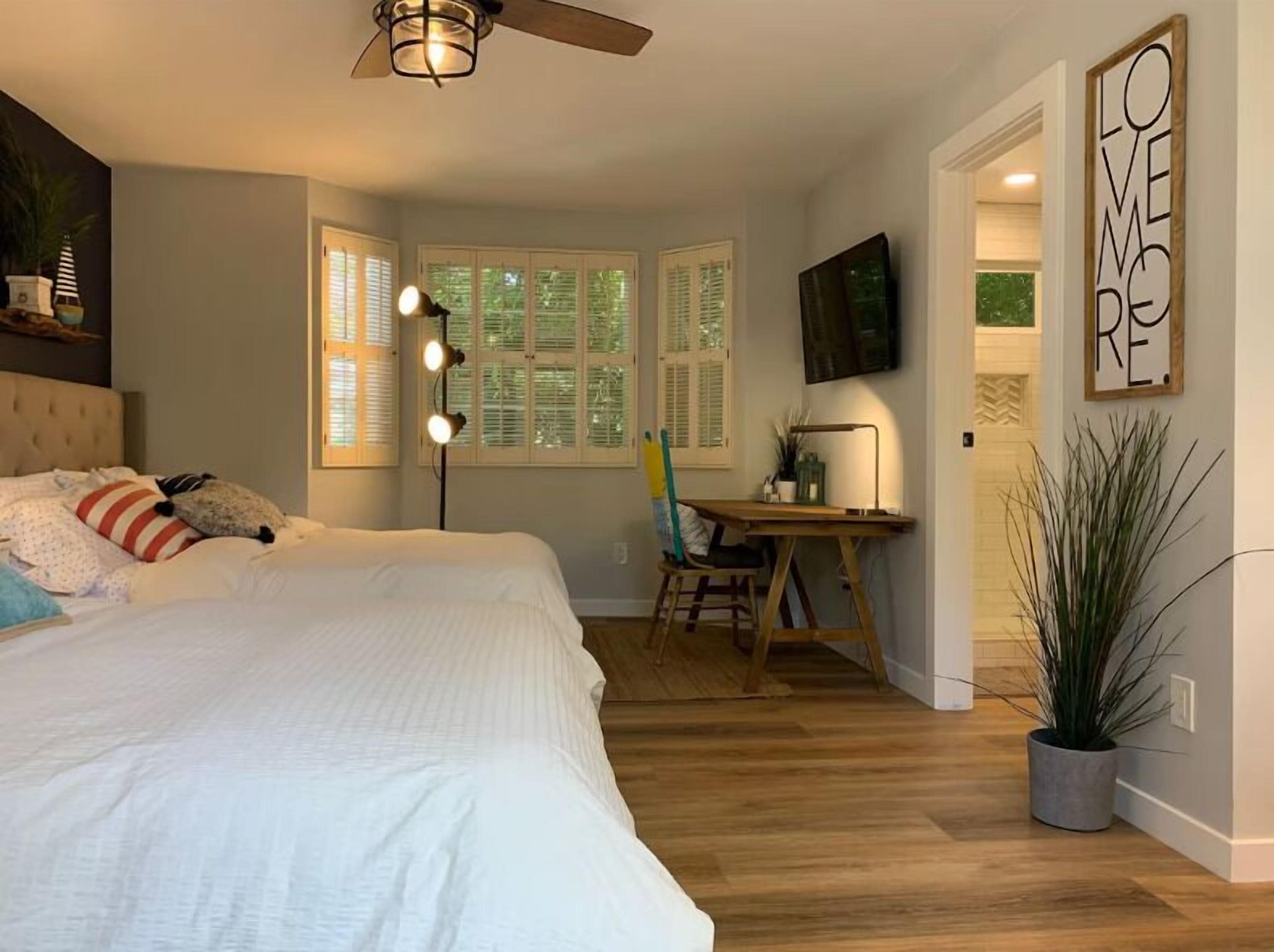 A bedroom with a bed, a desk, a chair, a ceiling fan, a TV, and a large window with shutters.