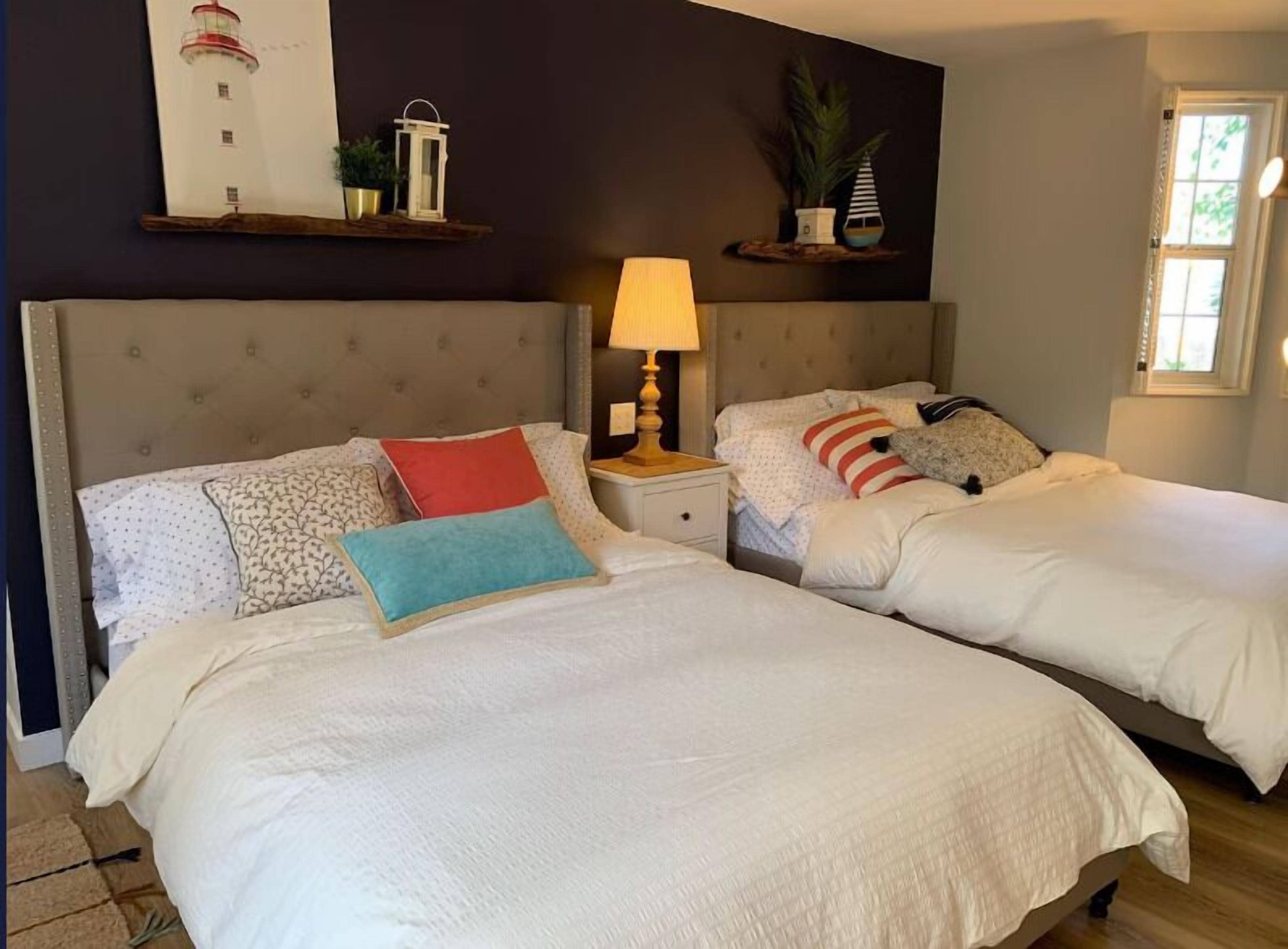A bedroom with two beds, a nightstand with a lamp, a wall-mounted shelf with decorative items, and a window.