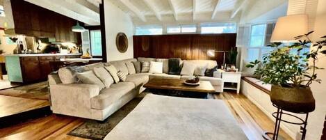 Una sala de estar moderna con un sofá modular, una mesa de centro y un otomán grande. Se ve un área de cocina al fondo.