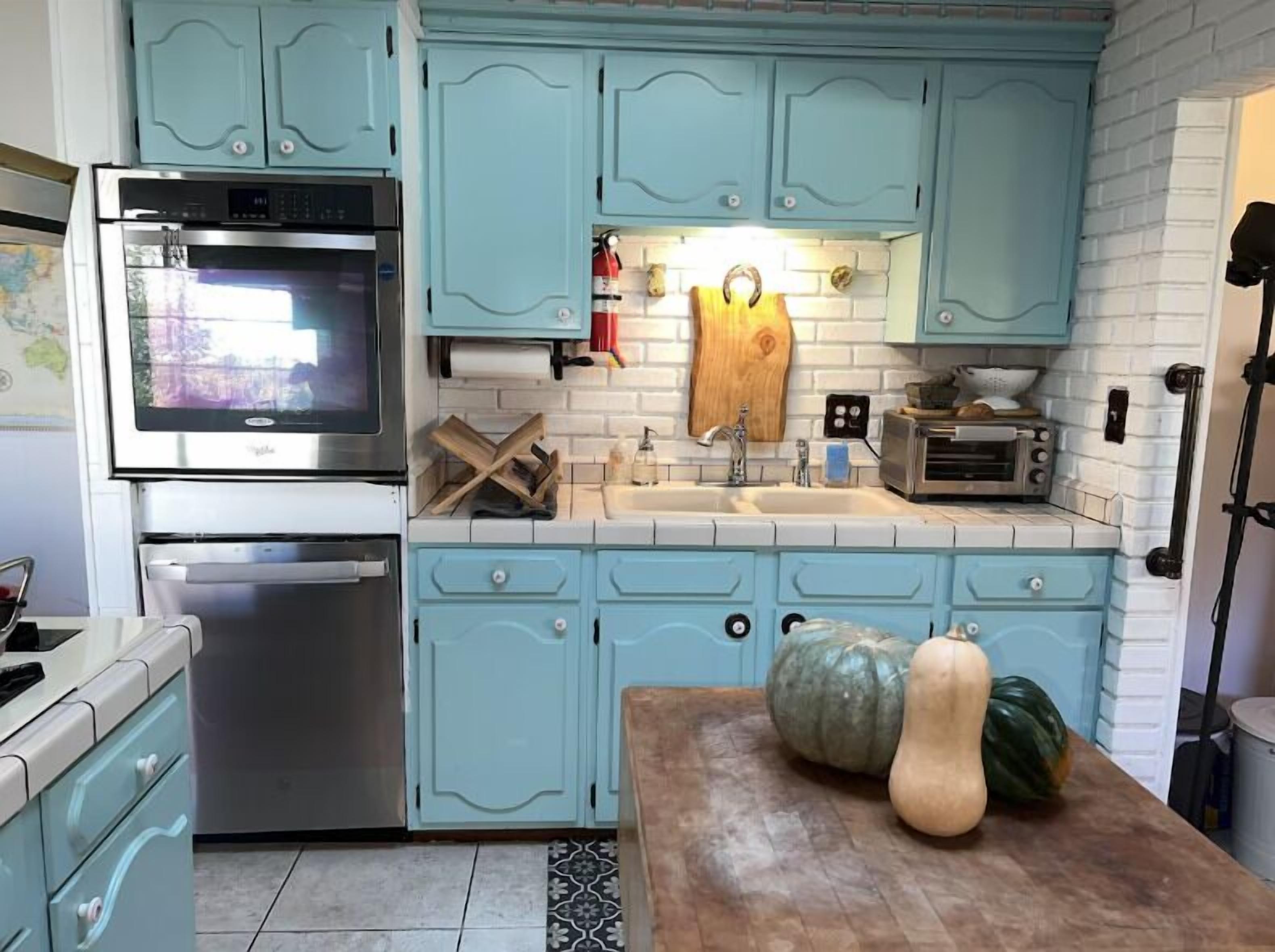 A kitchen with light blue cabinets, a stainless steel oven, a sink, and a wooden table with vegetables.