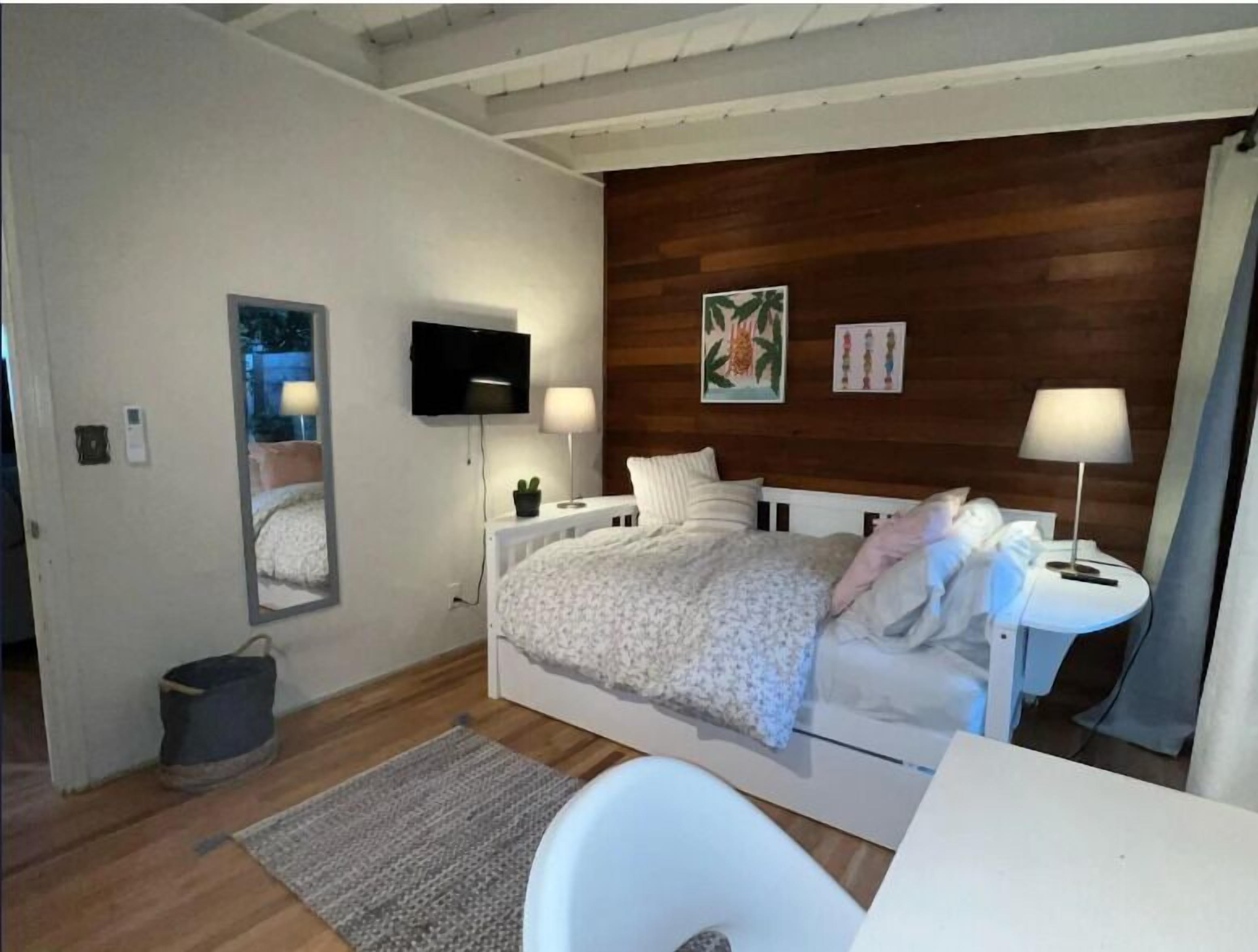 A bedroom with a wooden headboard, a bed with a white duvet, a bedside table with a lamp, a TV mounted on the wall, and a small potted plant.