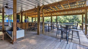 An outdoor patio with wooden tables and chairs, a covered seating area, and a view of greenery.