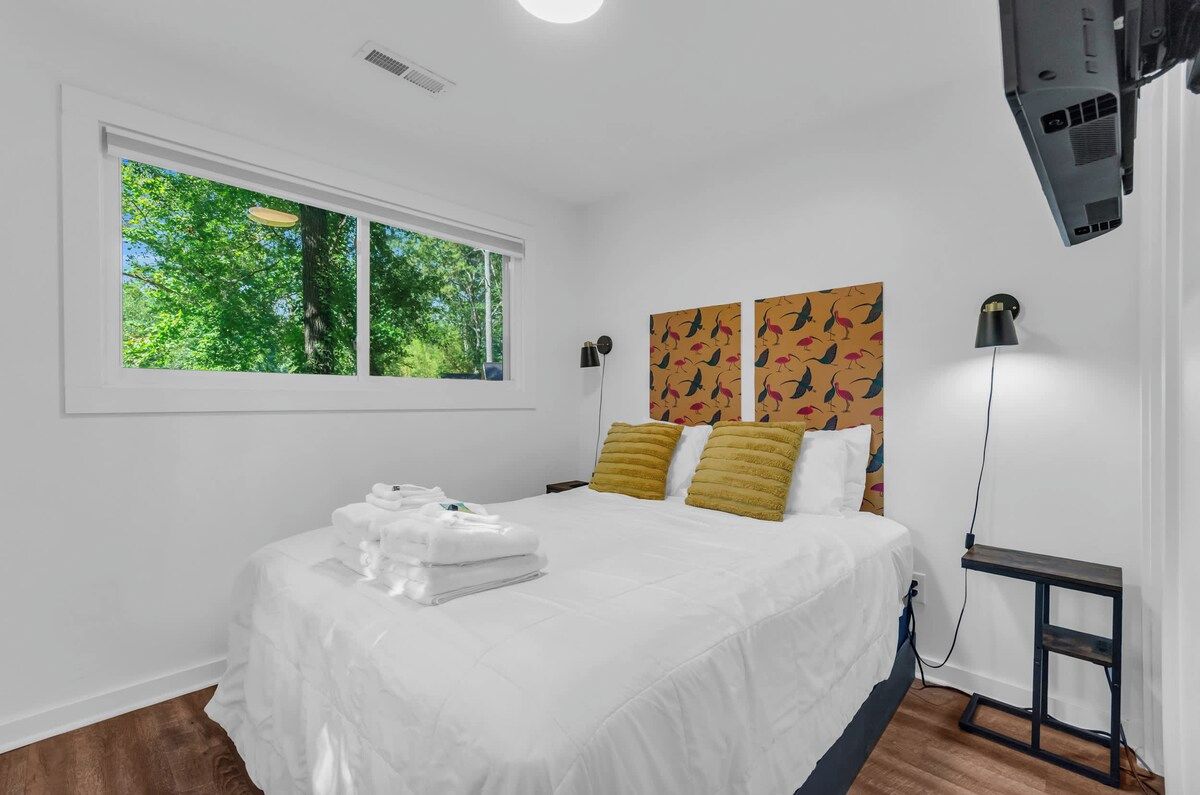 A hotel room with a bed, a window with a view of trees, a wall-mounted TV, and a bedside table.