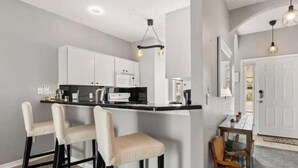 A modern kitchen with white cabinets, a black countertop, and three bar stools. There is a dining table with chairs and a small side table with a lamp. - Discover " a spacious 3-bedroom house  Enjoy a pool in the condo, fitness room, (Kissimmee)
