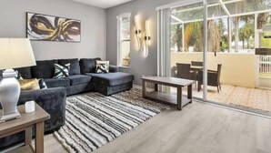 A modern living room with a grey sofa, a patterned rug, a ceiling fan, and a view of an outdoor patio area. - Discover " a spacious 3-bedroom house  Enjoy a pool in the condo, fitness room, (Kissimmee)