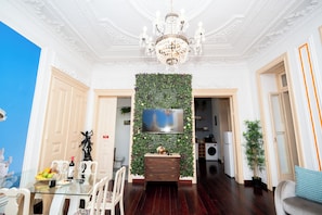 Ein Essbereich mit Glastisch, Stühlen, einer Chandelierleuchte, einem an der Wand montierten Fernseher und einer Wanddekoration mit Pflanzen.