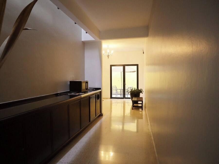 A hallway with a kitchen area on the left, a dining area with a table and chairs, and a view of the outdoors through a glass door.