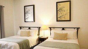 A hotel room with two beds, each with a white bedspread and a green throw blanket. There are two framed artworks on the walls, one above each bed. A bedside table with a lamp is between the beds, and a curtain is partially visible on the left. - Hotel Casa Kabii (Oaxaca)