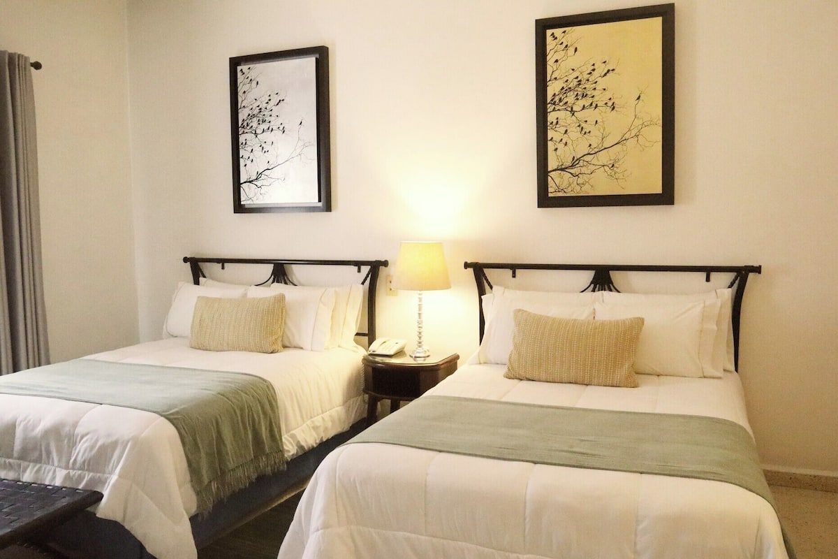 A hotel room with two beds, each with a white bedspread and a green throw blanket. There are two framed artworks on the walls, one above each bed. A bedside table with a lamp is between the beds, and a curtain is partially visible on the left.