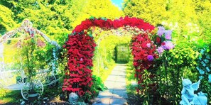 A garden pathway with a red floral archway, a white metal sculpture, and a statue of an angel. - Cozy 3BR Villa · Big garden rose· photos for festival ,Near Beach, breakfast  (Surrey)