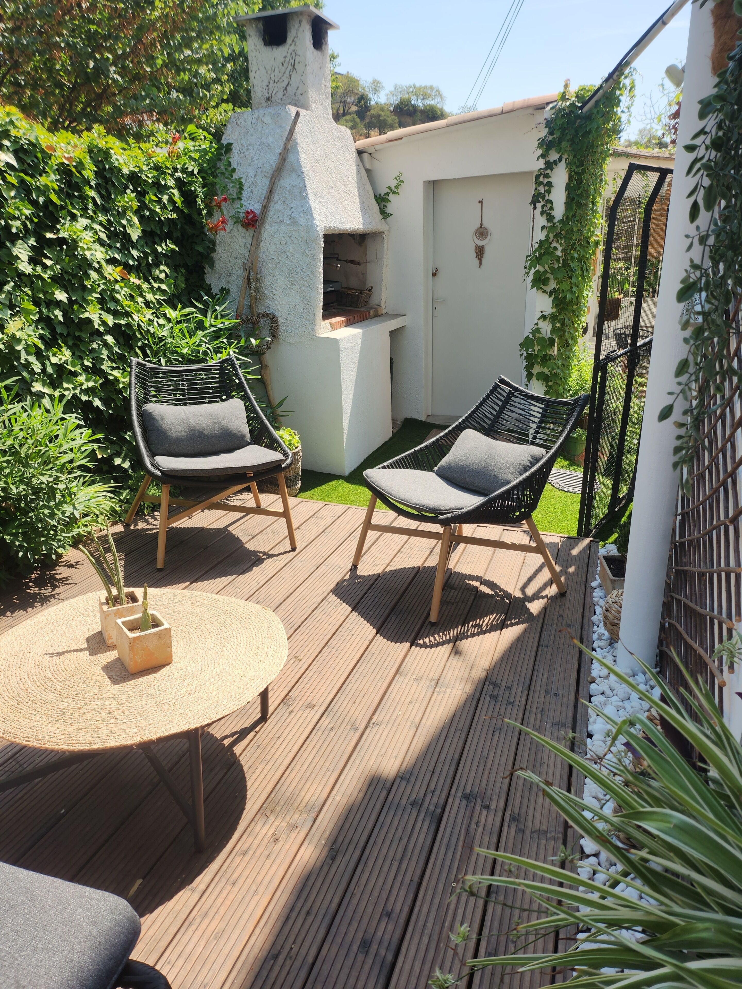 A wooden deck with two black wicker chairs and a round table, surrounded by greenery and a white wall.
