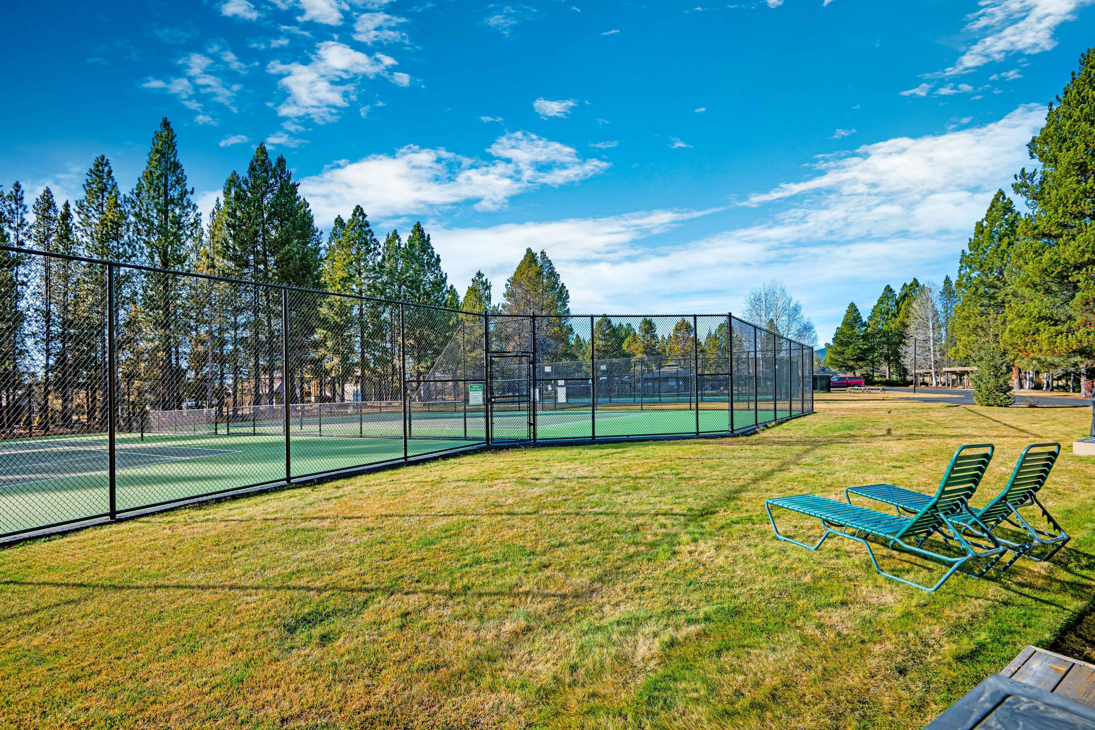 A tennis court with a fence, green lawn chairs, and trees in the background.