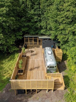 Eine Holzterrasse mit einem Wohnmobil, einer überdachten Sitzgelegenheit und einem Gebäude mit großen Fenstern.