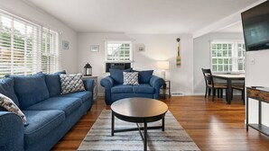 A modern living room with a blue sofa, a round coffee table, and a dining area with a table and chairs. - Modern Falmouth Home Close to Water and Town (Falmouth)