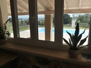 Vista a través de una ventana con un alero de piedra, una piscina y follaje.