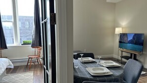 A hotel room with a bed, a window with curtains, a dining area with a table set for two, and a TV mounted on the wall.