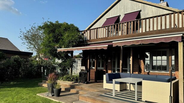 Een tweelaags huis met een overdekt terras, buitenmeubels en een groot raam met uitzicht op de tuin.