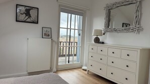 Een slaapkamer met een groot raam, een witte dressoir, een spiegel en een ingelijste foto aan de muur.