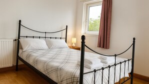 Una cama bien tendida con una colcha escocesa, una mesita de noche con una lámpara, y una ventana con cortinas.