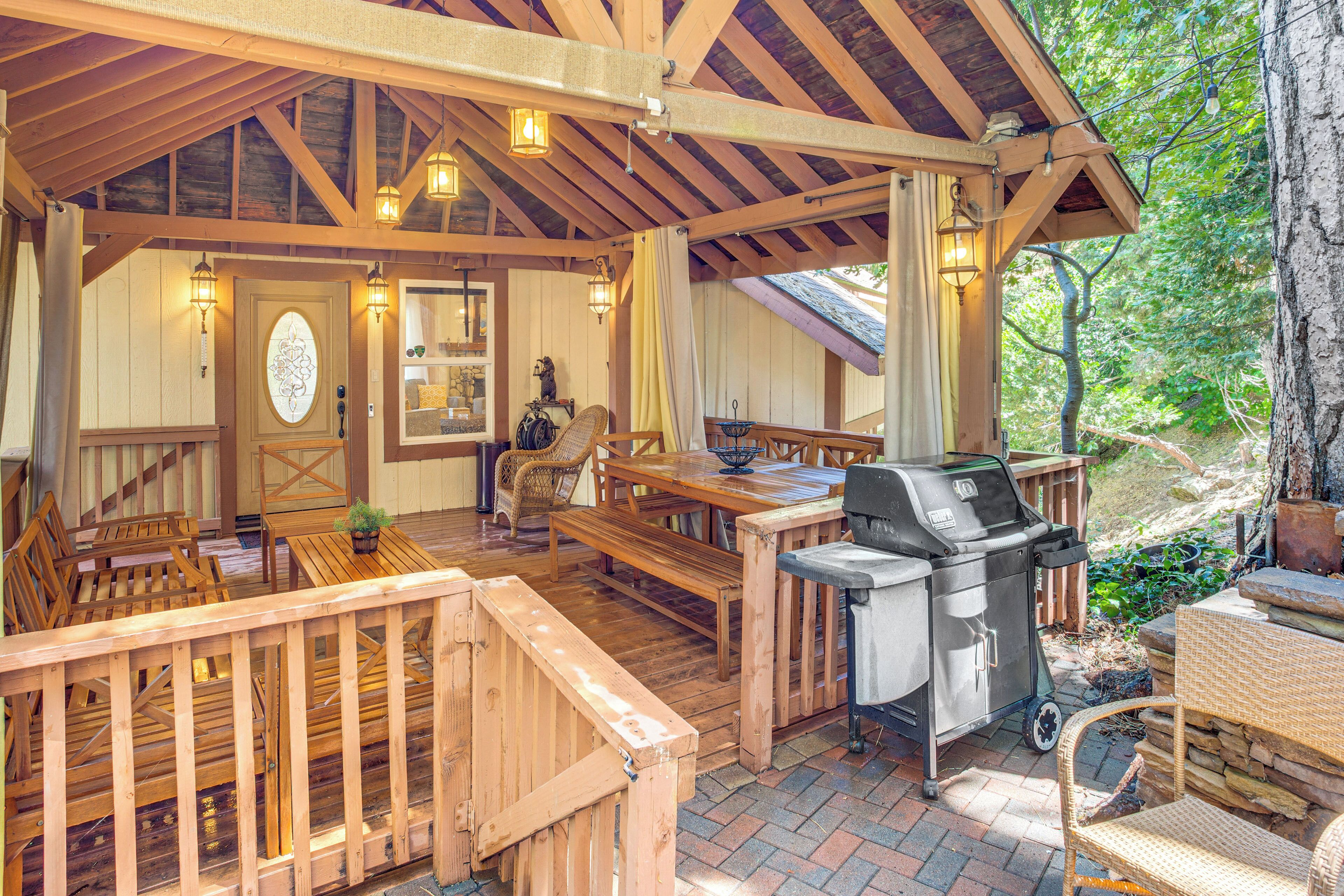 Un patio techado al aire libre con mobiliario de madera, una parrilla y vistas del bosque circundante.