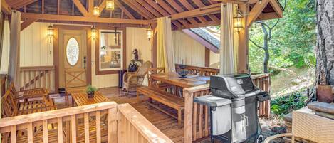 Un patio techado al aire libre con mobiliario de madera, una parrilla y vistas del bosque circundante.