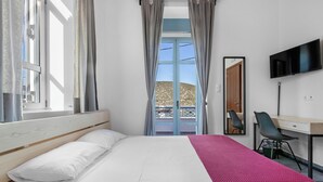 Quarto com cama, escrivaninha, cadeira, televisão e varanda com vista para uma colina.