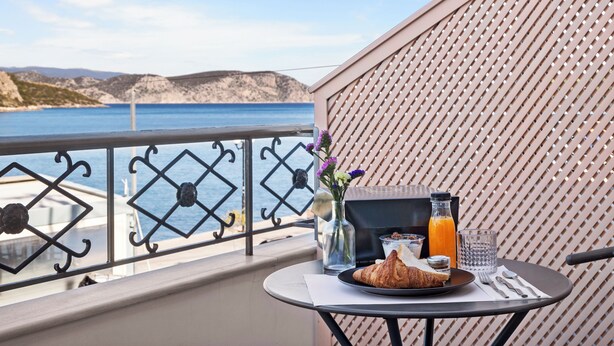 Varanda com mesa posta para o café da manhã, um vaso com flores e vista para o mar e montanhas.