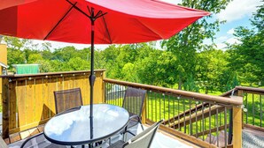 Una terraza de madera con una mesa y sillas de vidrio, un paraguas rojo y vistas a un césped verde y árboles.