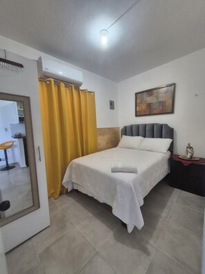 Un dormitorio con cama, mesita de noche, un cuadro y baño visible a través de una puerta abierta.