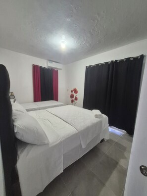 Una habitación con cama, aire acondicionado de pared, ventana con cortinas negras y decoración floral roja en la pared.