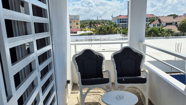 A balcony with two black cushioned chairs and a small white table, white railings, patterned floor tiles, and a view of buildings and palm trees.
