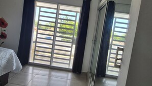 A room with a large sliding glass door leading to an outdoor area, a bed with a white cover, and a vase with a single red flower.