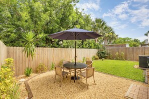 Une terrasse à l’arrière de la maison, avec une table ronde et des chaises, entourée d’une clôture en bois, et protégée par un grand parasol.