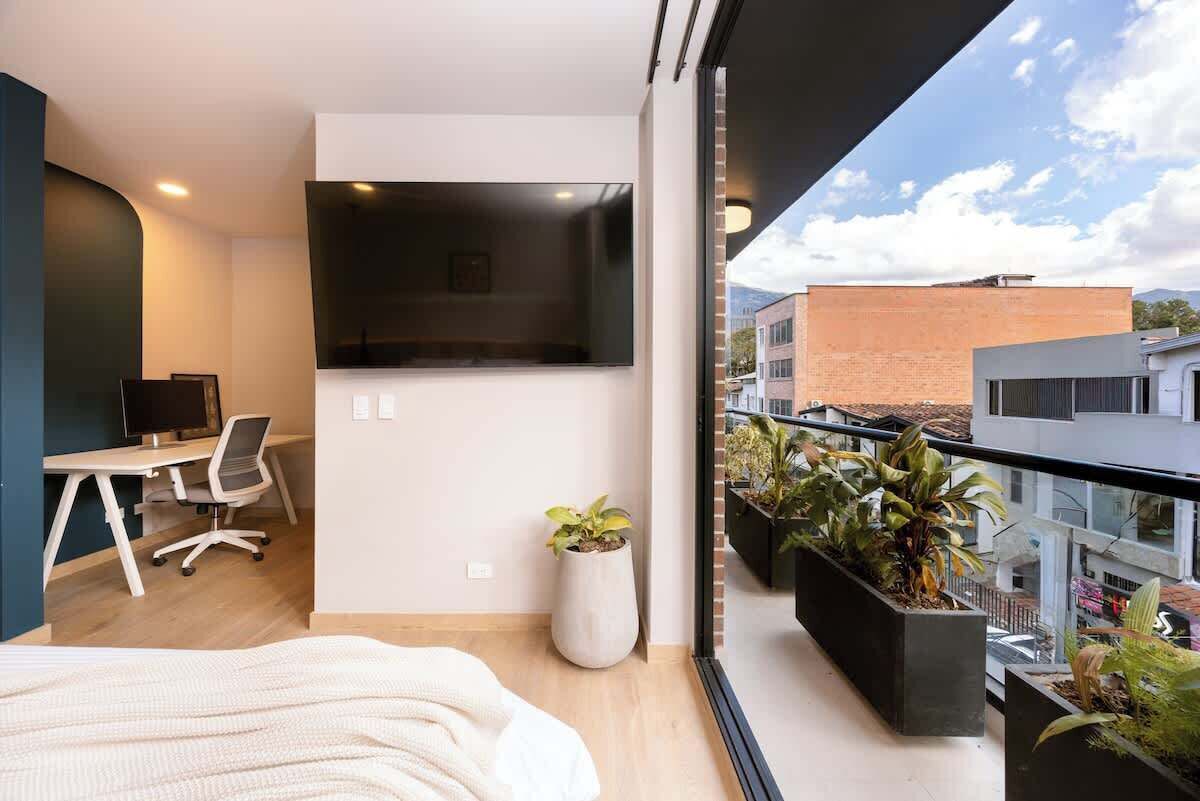 Quarto moderno com cama, escrivaninha, cadeira, televisão e varanda com plantas.