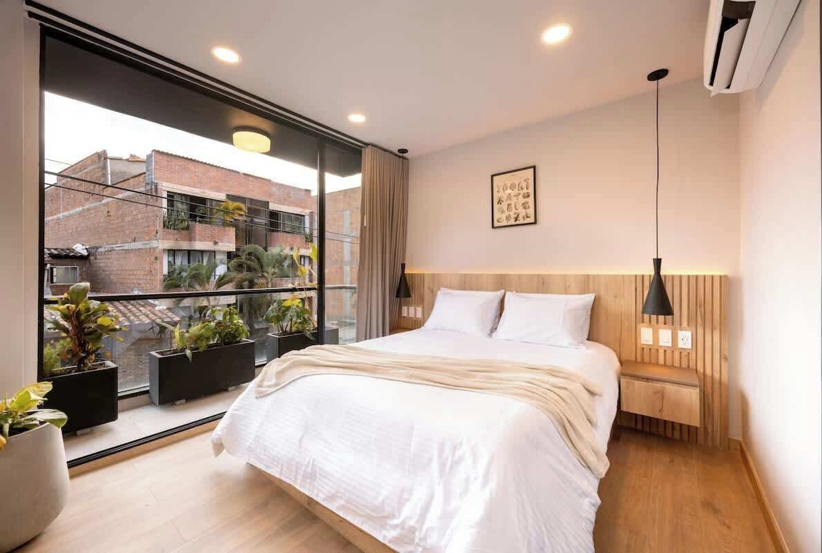 A modern bedroom with a large bed, a wooden headboard, a bedside table with a lamp, and a view of buildings through a large window.