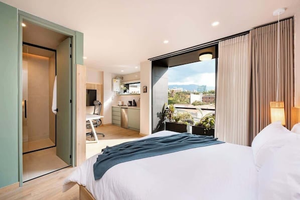 A modern hotel room with a large bed, a kitchenette, and a balcony with a city view.