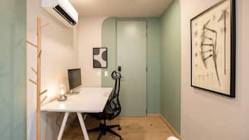 Um pequeno escritório com uma escrivaninha branca, um computador, uma cadeira, um ar condicionado e um quadro abstrato na parede.