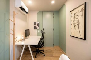 A small office with a white desk, a computer, a chair, an air conditioner, and a framed abstract painting on the wall.