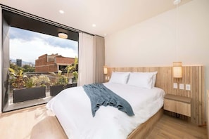 Une chambre d’hôtel moderne avec un grand lit, une tête de lit en bois, une table de chevet et une vue sur la ville grâce à de grandes fenêtres.