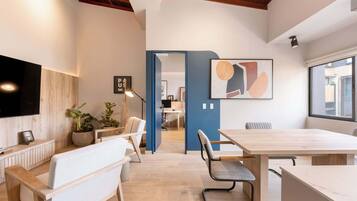 Un espacio de oficina moderno con un banco de madera, un escritorio y una mesa grande. Hay plantas en macetas y un cuadro en la pared.