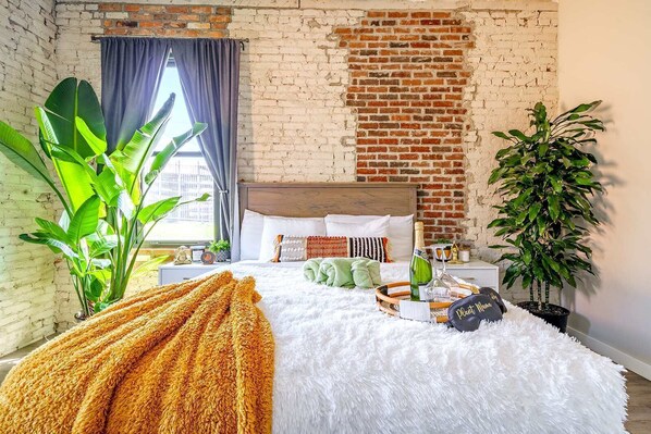 Un dormitorio con pared de ladrillo, cama con ropa de cama blanca, una planta grande y una ventana con cortinas.