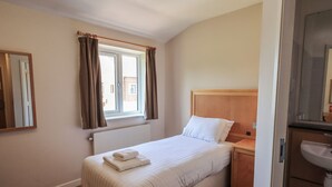 A hotel room with a single bed, a nightstand, a window with curtains, a radiator, and a bathroom with a sink. - Jubilee Single Bedroom Ground-floor (Penrith)