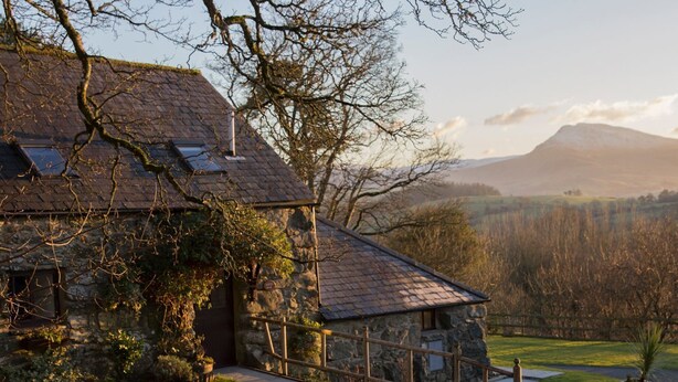 A stone house with a sloped roof, surrounded by a well-maintained lawn and a view of a mountain range. - Cyffdy Cottage - Tegid (Bala)