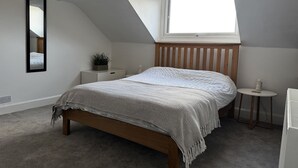 A bedroom with a sloped ceiling, a bed with a patterned blanket, a bedside table with a plant, and a small round table with a lamp. - Mostyn View (Wirral)