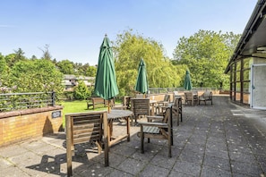 Een terras buiten met houten tafels en stoelen, groene parasols en een bakstenen muur.