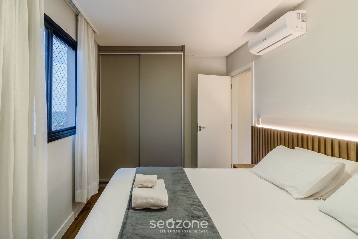 A hotel room with a bed, a bedside table, a window with curtains, a wardrobe, and an air conditioning unit.