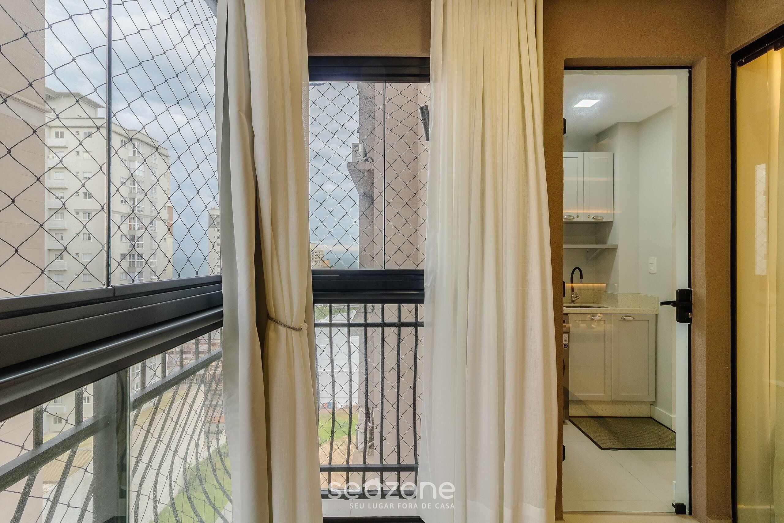 Vista desde un balcón con una valla de alambre, una puerta que da a una cocina y un edificio al fondo.