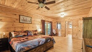 Une chambre à coucher dans une cabane en bois, avec un lit, un ventilateur de plafond et une porte donnant sur une autre pièce.