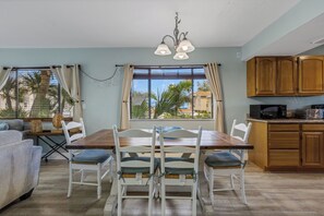 Un coin repas avec une table en bois et des chaises blanches, une cuisine avec des meubles en bois et une vue sur les palmiers par les fenêtres.